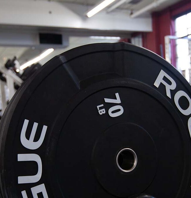 Fitness plateau in NJ: Close-up of a 70LB Rogue Plate with a glimpse of the gym in the background, photo taken during the day.