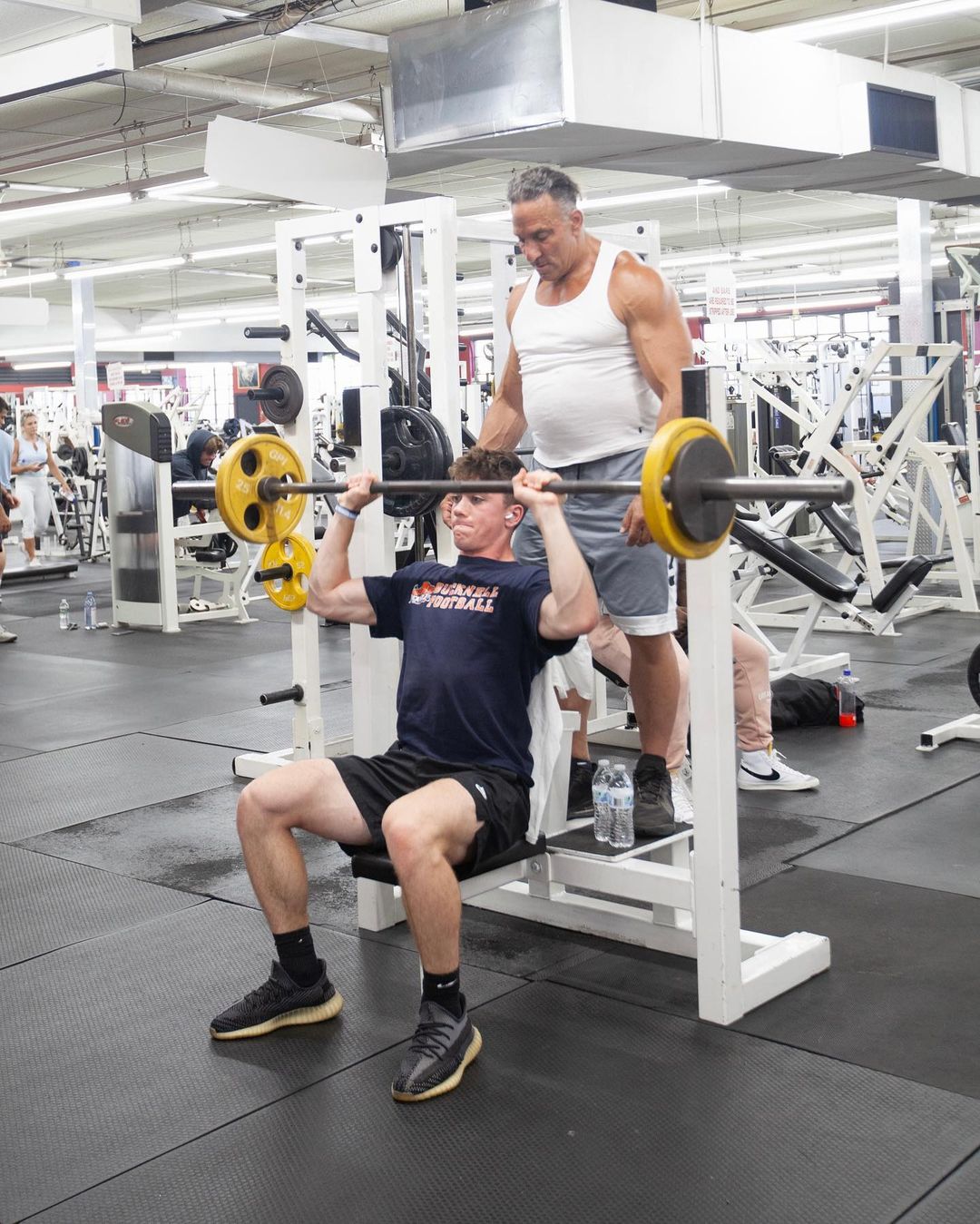 Gym Etiquette NJ - Man spotting young boy during barbell shoulder press in a gym, embodying gym etiquette and support.